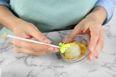 Young woman putting cake pop into yellow sprinkles at white marble table, closeup