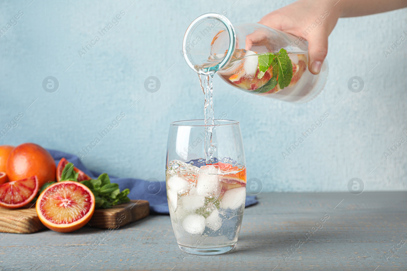 Photo of Woman pouring refreshing drink with sicilian orange from bottle into glass at grey wooden table, closeup