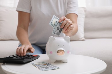 Woman with calculator putting money into piggy bank at table indoors, closeup