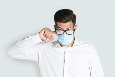 Man wiping foggy glasses caused by wearing medical mask on light background