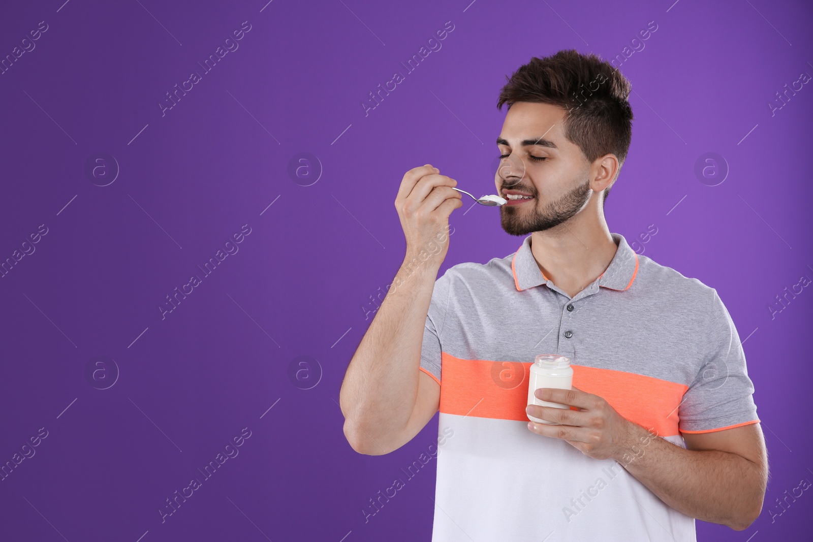 Photo of Happy young man eating tasty yogurt on purple background. Space for text