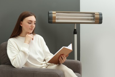 Photo of Young woman reading book on armchair near electric infrared heater at home. Space for text