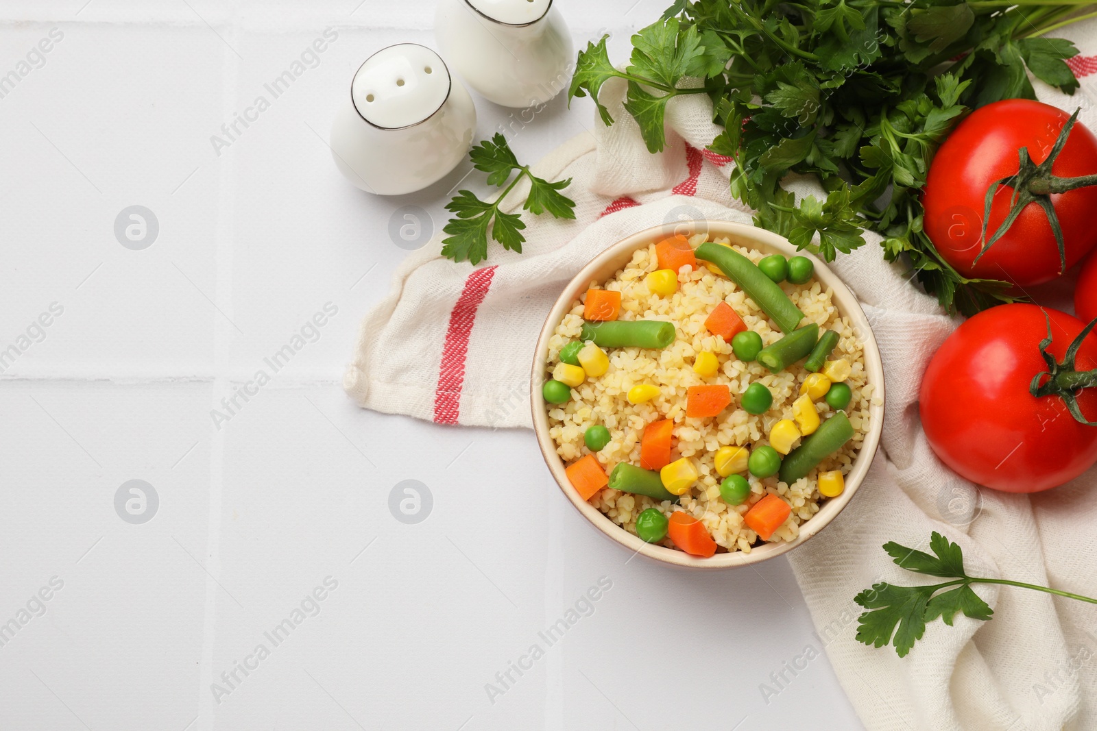 Photo of Delicious bulgur with vegetables in bowl, tomatoes and parsley on white tiled table, flat lay. Space for text