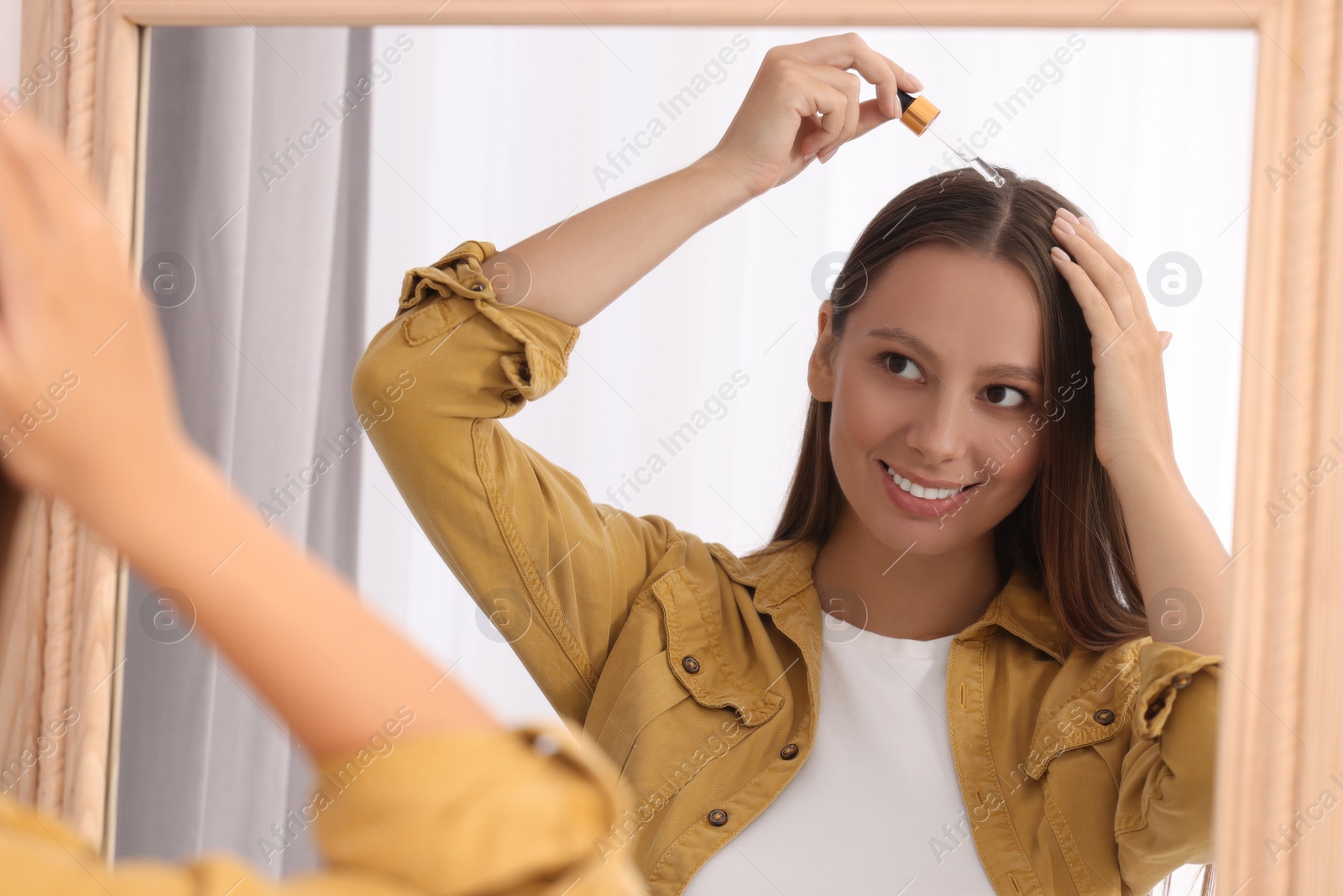 Photo of Beautiful woman applying serum onto hair near mirror indoors