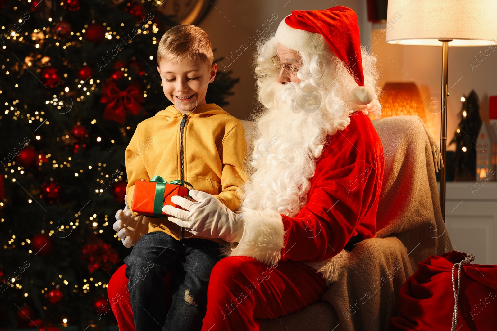 Photo of Merry Christmas. Smiling little boy sitting on Santa's knee and holding gift at home