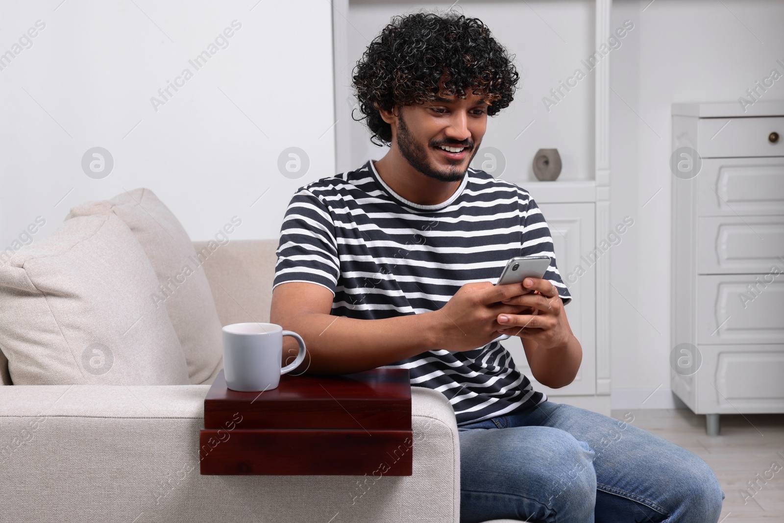 Photo of Happy man using smartphone on sofa with wooden armrest table at home