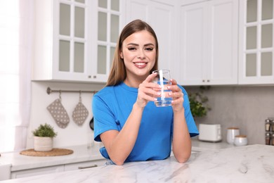 Photo of Happy woman with glass of fresh water at white marble table in kitchen