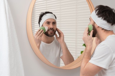 Photo of Emotional man with headband washing his face using sponge near mirror in bathroom