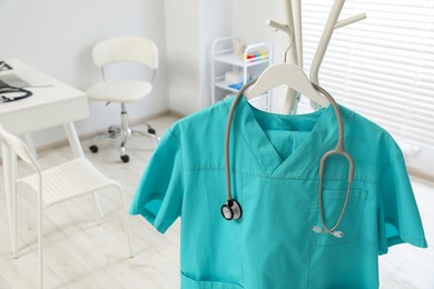 Turquoise medical uniform and stethoscope on rack in clinic, closeup