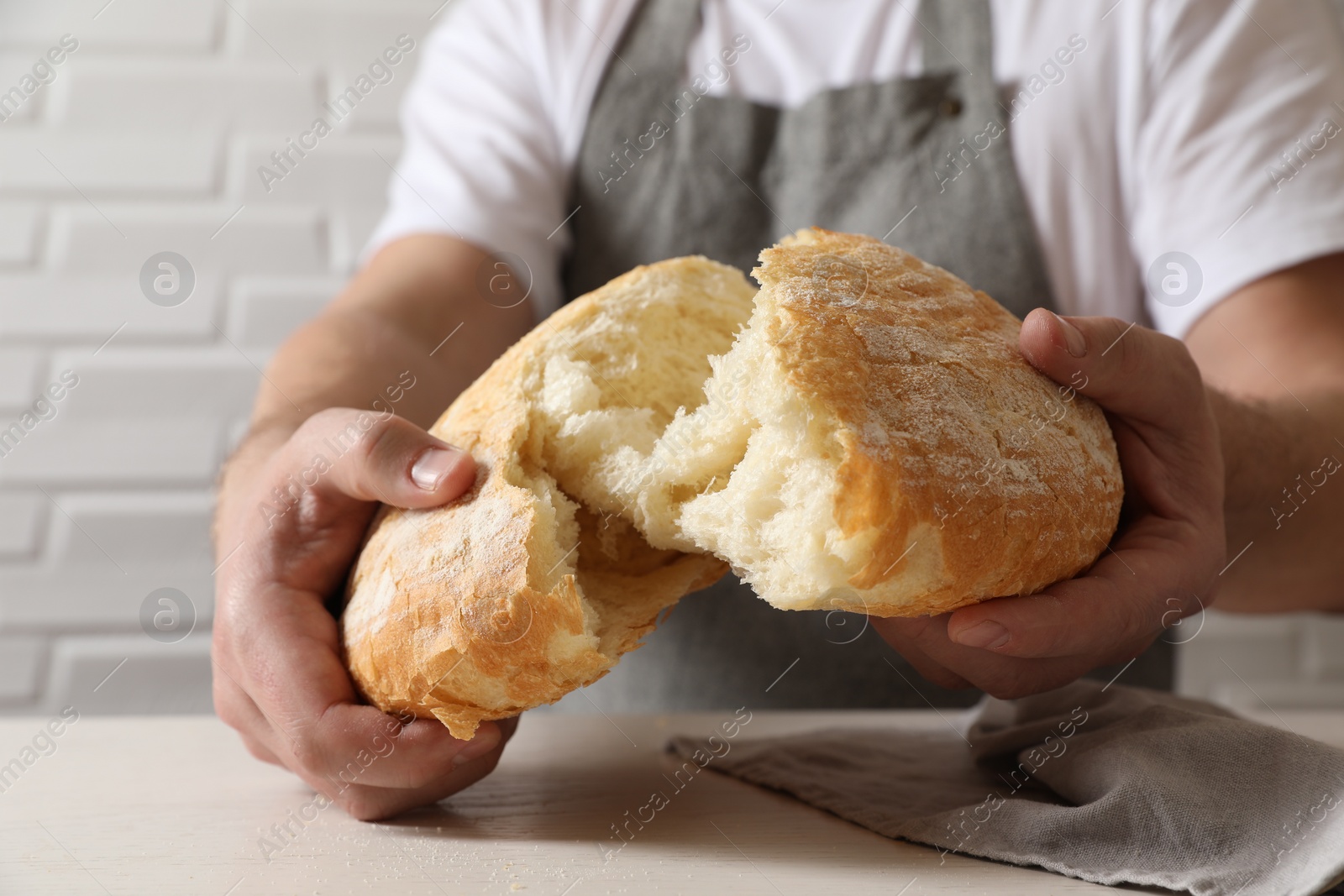 Photo of Man breaking loaf of fresh bread at white table near brick wall, closeup