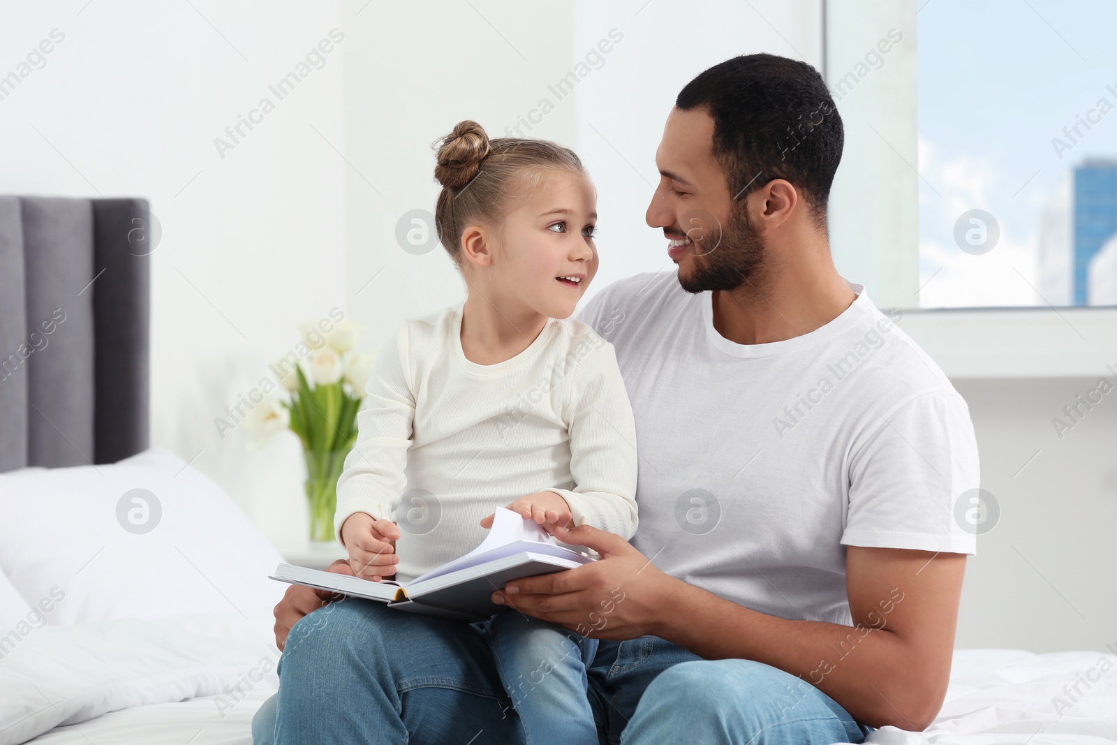 Photo of Little girl with her father reading book together on bed at home. International family