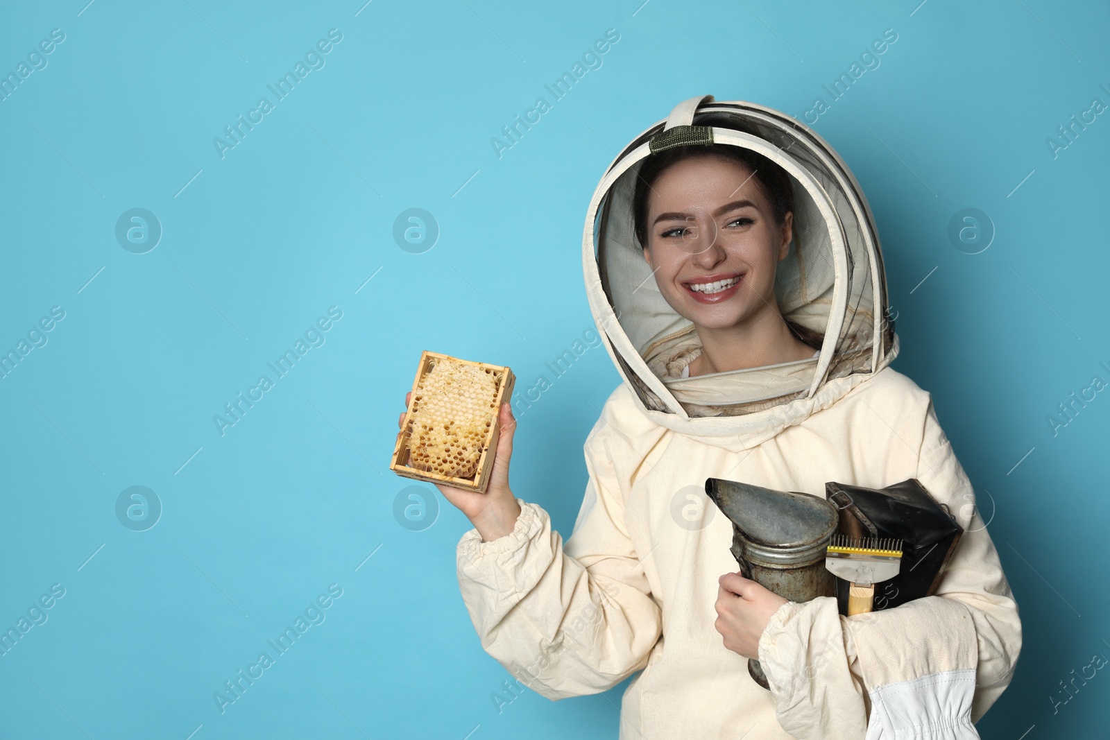 Photo of Beekeeper in uniform holding smokepot and hive frame with honeycomb on light blue background. Space for text