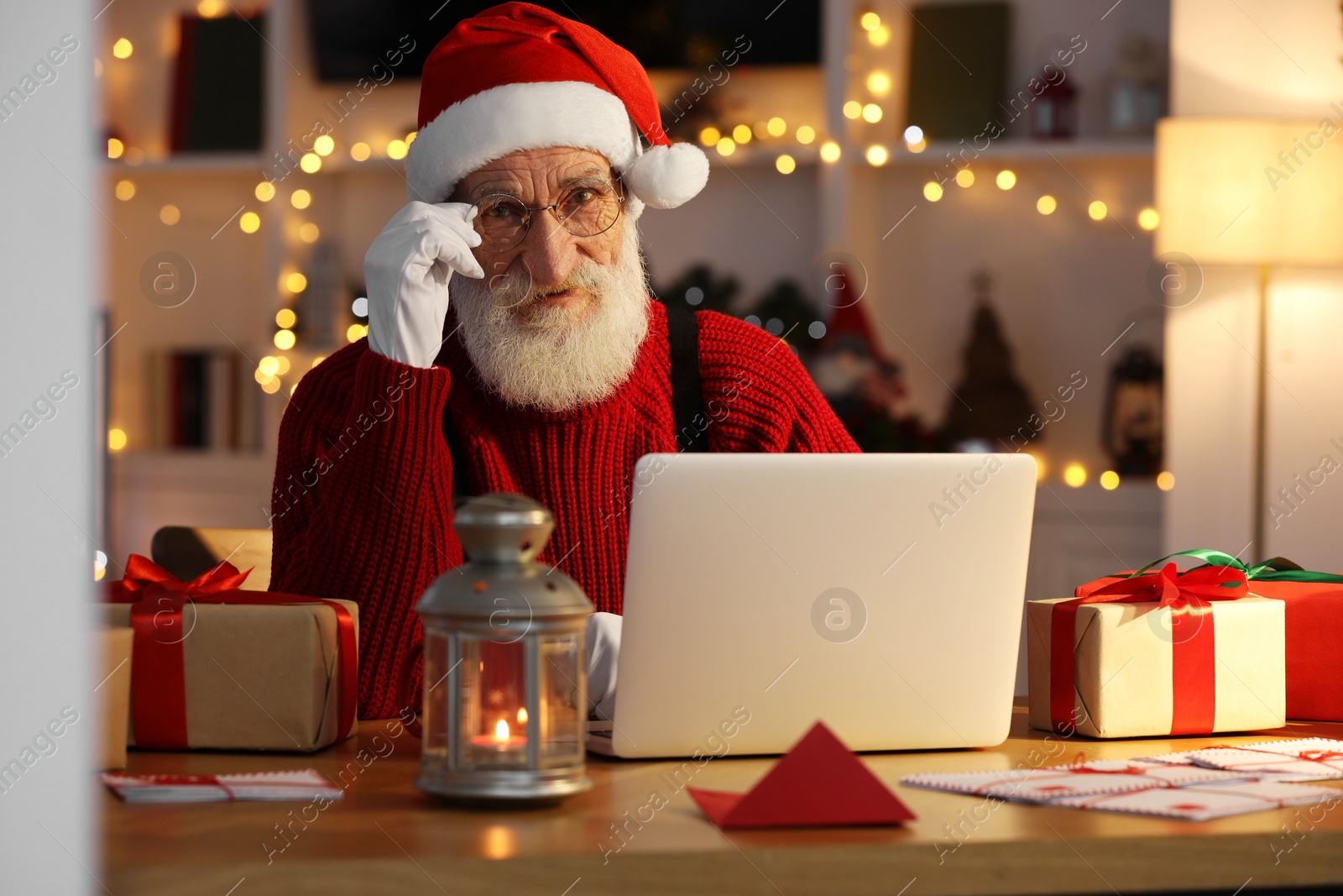 Photo of Santa Claus using laptop at his workplace in room decorated for Christmas