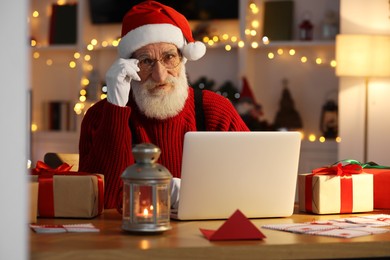 Photo of Santa Claus using laptop at his workplace in room decorated for Christmas