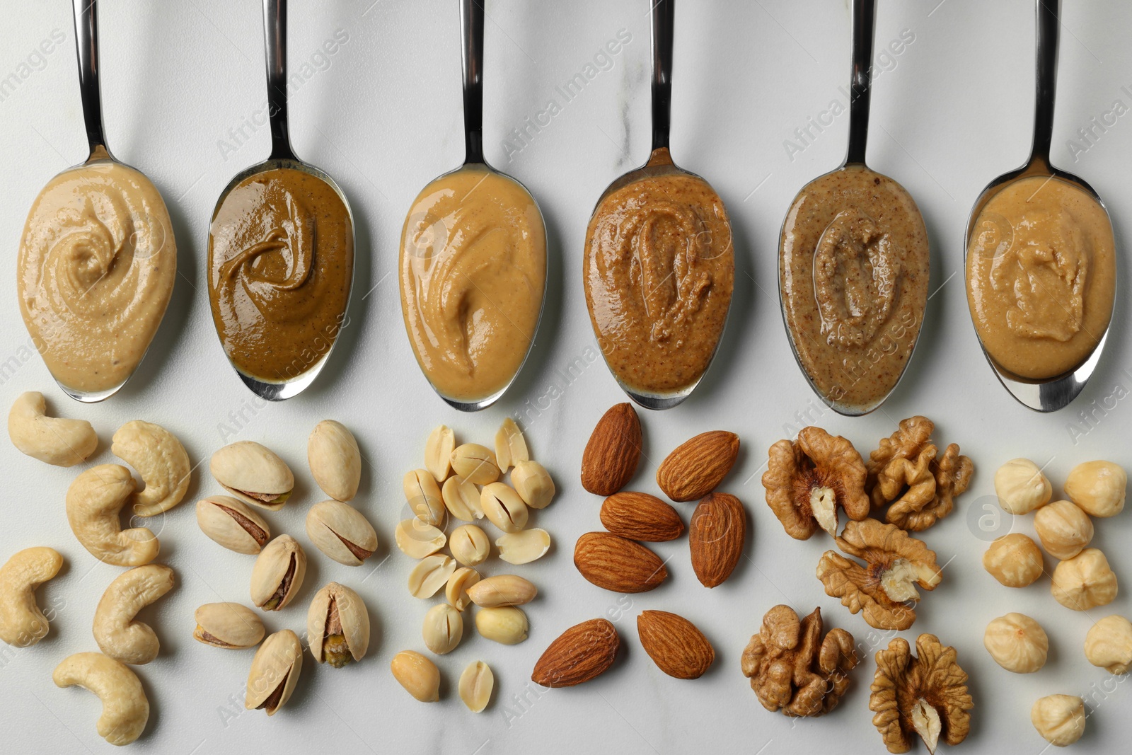 Photo of Tasty nut butters in spoons and raw nuts on white marble table, flat lay