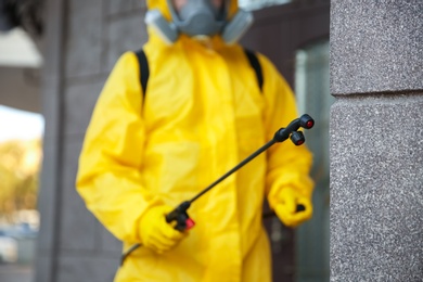 Photo of Person in hazmat suit disinfecting building wall outdoors, focus on sprayer. Surface treatment during coronavirus pandemic