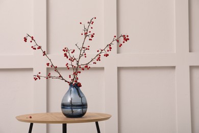 Photo of Hawthorn branches with red berries in vase on wooden table near white wall indoors, space for text. Interior element