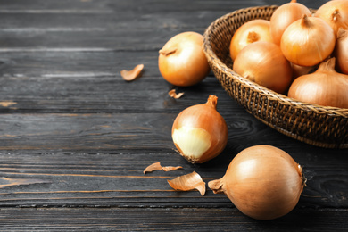 Ripe onion bulbs and basket on black wooden table, closeup. Space for text