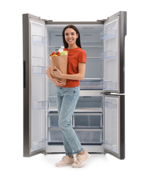 Photo of Young woman with bag of groceries near empty refrigerator on white background