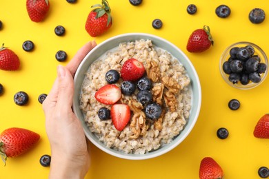 Photo of Woman holding bowl of tasty oatmeal with strawberries, blueberries and walnuts on yellow background, top view