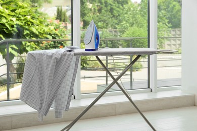 Photo of Modern iron and clean checkered shirt on board near window indoors