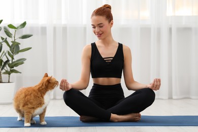 Beautiful woman with cute red cat practicing yoga on mat at home