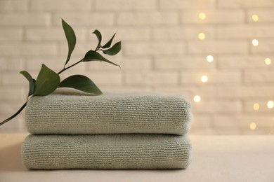 Stacked soft towels and green branch on white table near brick wall indoors, space for text