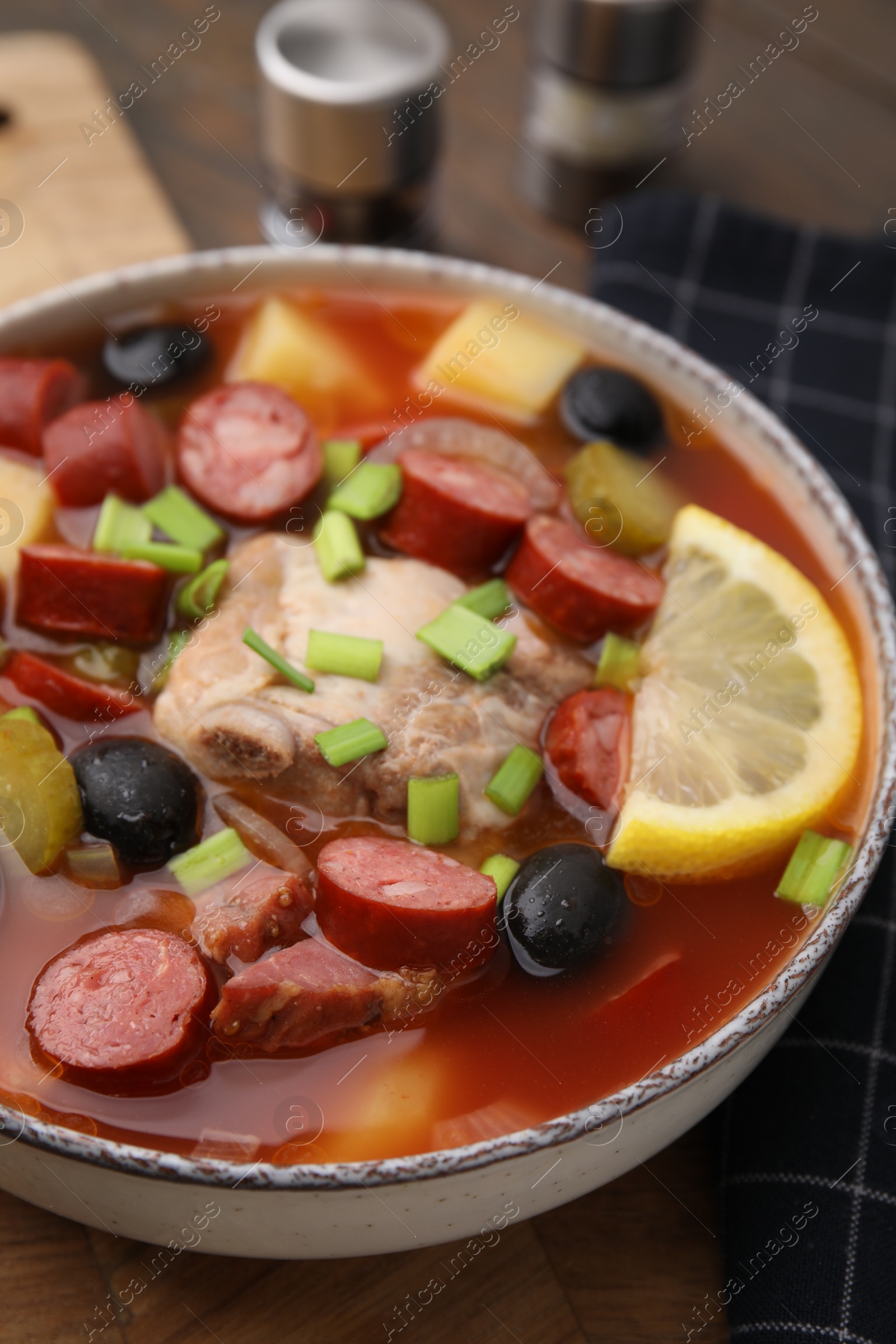 Photo of Meat solyanka soup with thin dry smoked sausages in bowl on wooden board, closeup