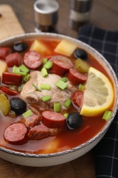 Photo of Meat solyanka soup with thin dry smoked sausages in bowl on wooden board, closeup