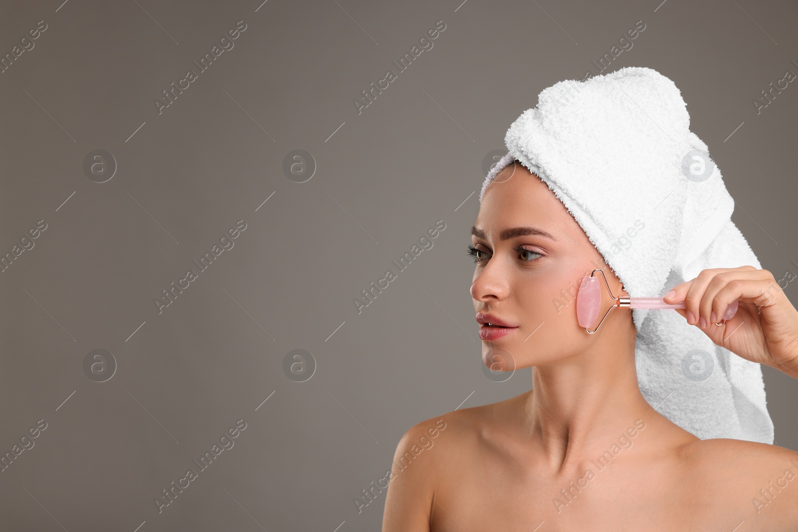 Photo of Young woman massaging her face with rose quartz roller on grey background, space for text