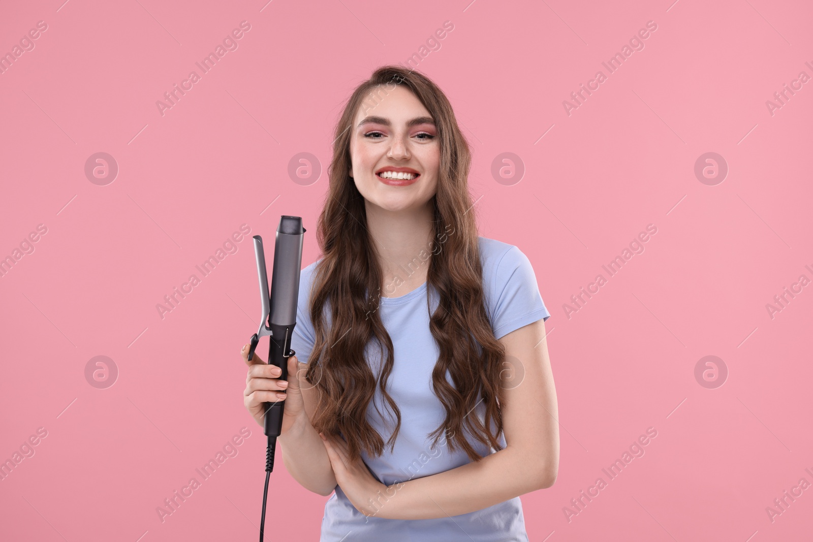 Photo of Happy young woman with beautiful hair holding curling iron on pink background