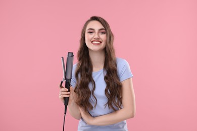 Happy young woman with beautiful hair holding curling iron on pink background