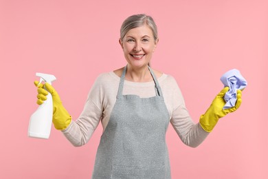 Happy housewife with spray bottle and rag on pink background