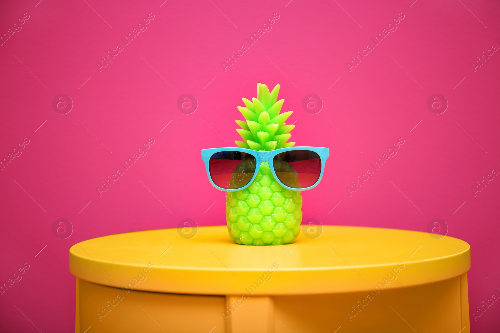 Photo of Stylish pineapple candle with sunglasses on table against color background. Home decorating idea