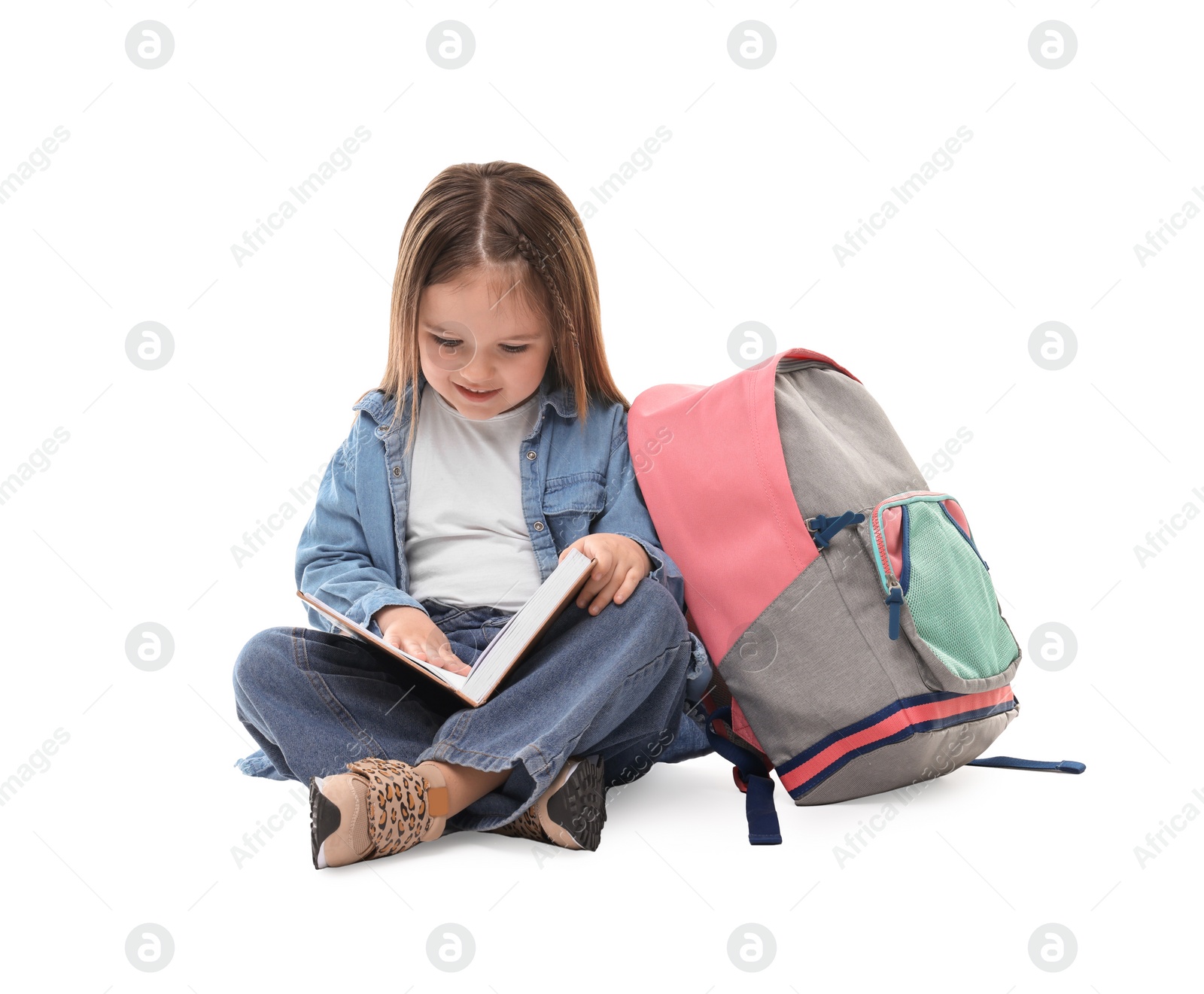 Photo of Cute little girl reading book near backpack on white background