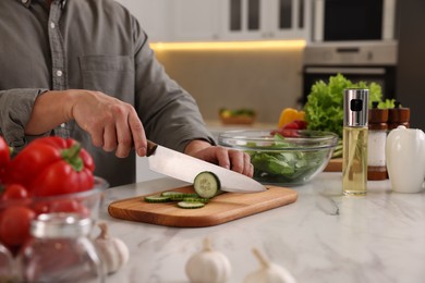 Photo of Cooking process. Man cutting fresh cucumber at white marble countertop in kitchen, closeup