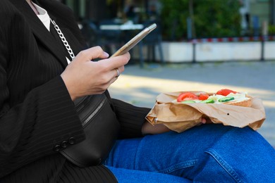 Photo of Woman holding tasty sandwich with vegetables and using phone outdoors, closeup. Street food