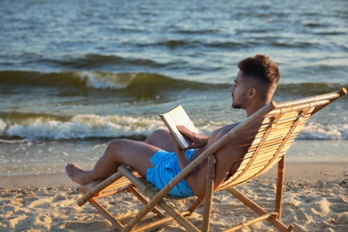 Young man reading book on sandy beach near sea