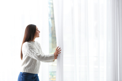 Young woman opening window curtains at home