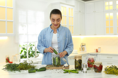 Woman putting cucumber into pickling jar at table in kitchen