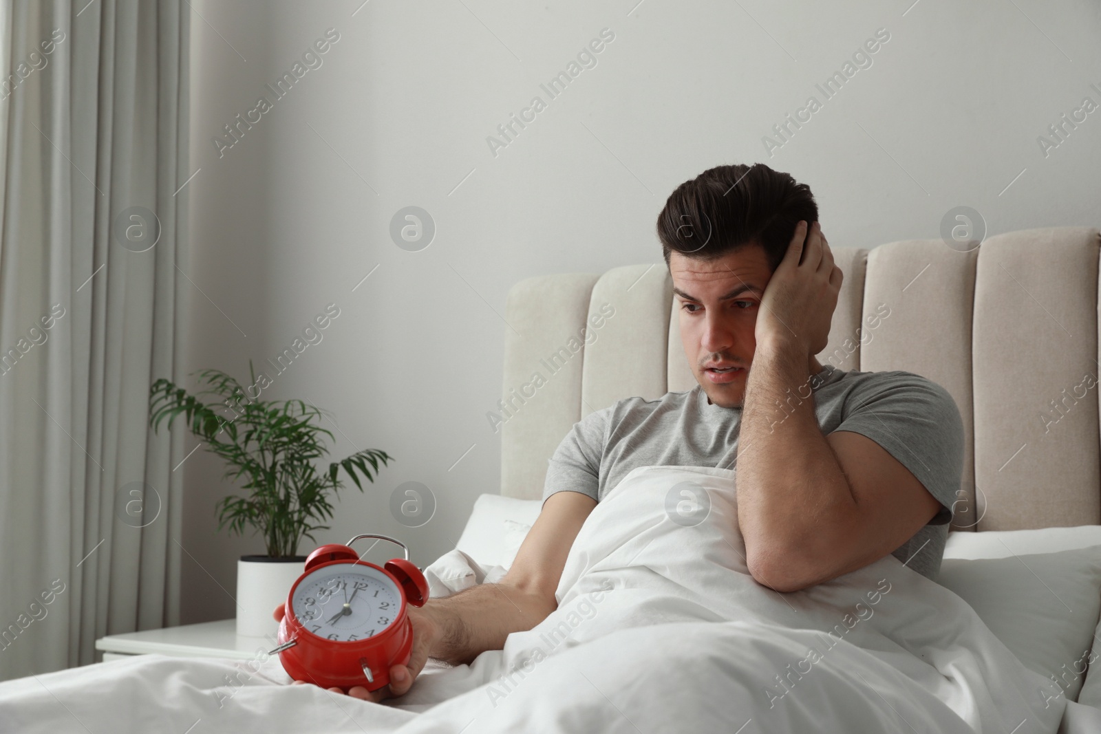 Photo of Emotional man with alarm clock in bed. Being late because of oversleeping