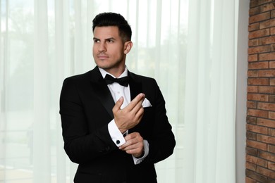 Handsome stylish man putting on cufflink near window indoors