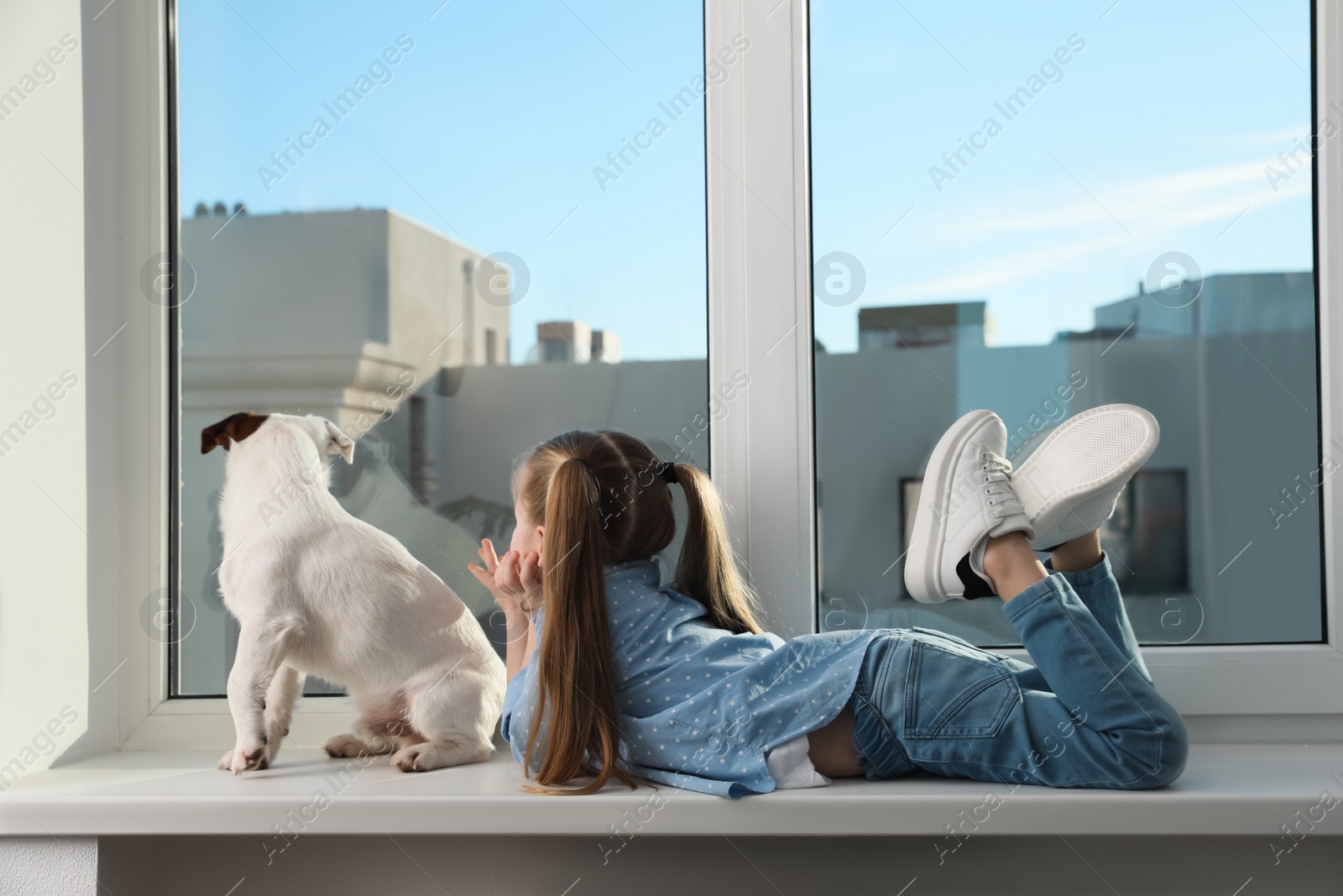 Photo of Cute little girl with her dog on window sill indoors. Childhood pet