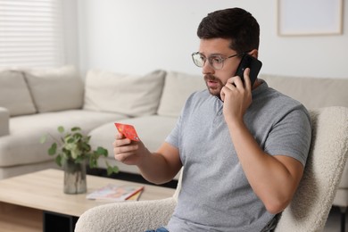 Upset man with credit card talking on smartphone in armchair at home, space for text. Be careful - fraud