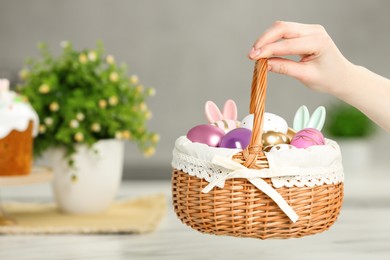 Photo of Woman holding Easter basket with painted eggs and decorations near white table, closeup. Space for text