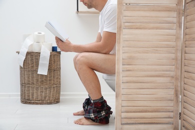 Young man with book sitting on toilet bowl in bathroom