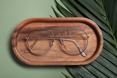 Stylish pair of glasses, tray and palm leaf on light green background, flat lay