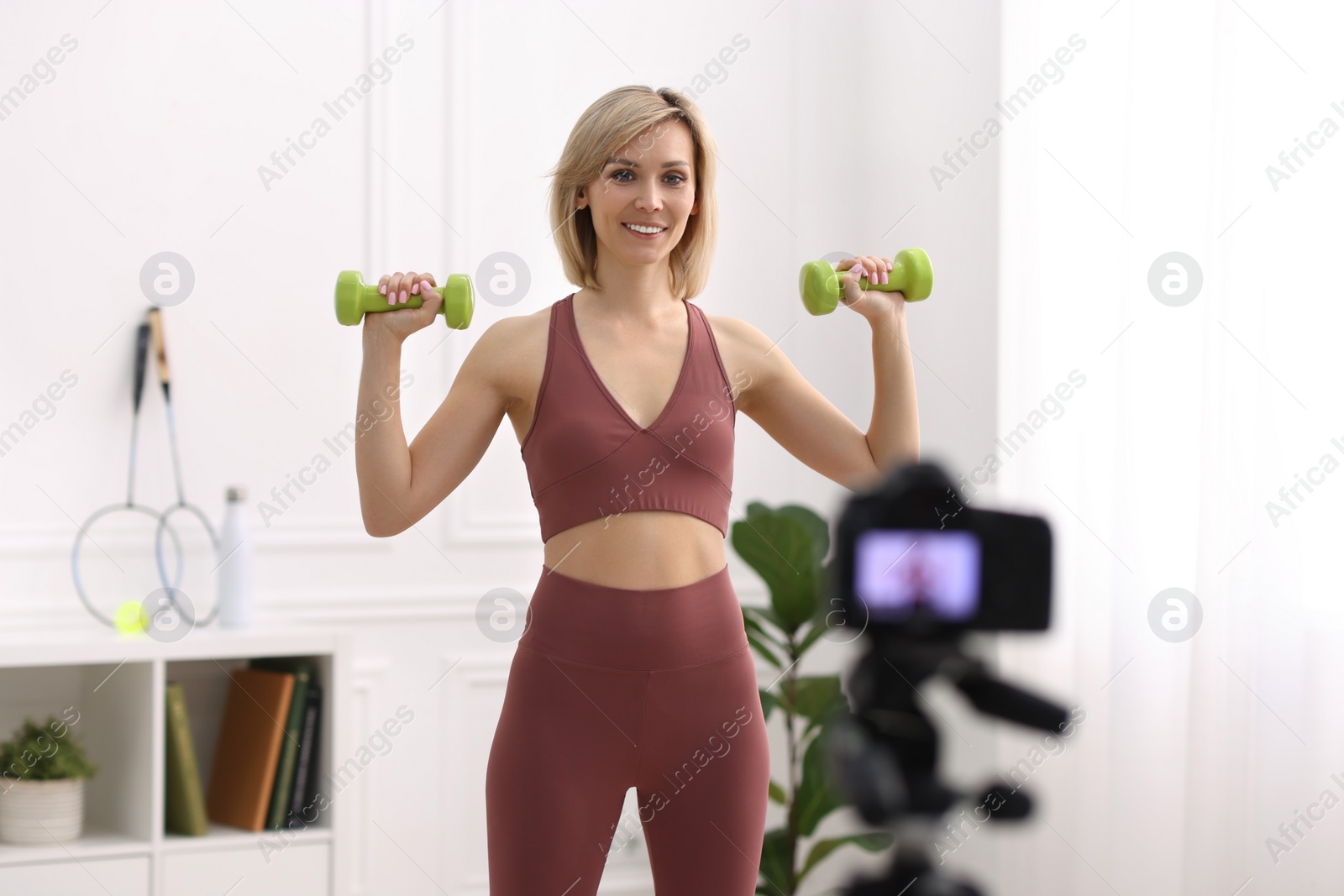 Photo of Smiling sports blogger working out with dumbbells while recording fitness lesson at home