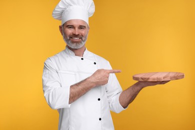 Photo of Happy chef in uniform pointing at wooden board on orange background