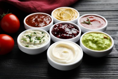 Photo of Different tasty sauces in bowls and tomatoes on black wooden table, closeup